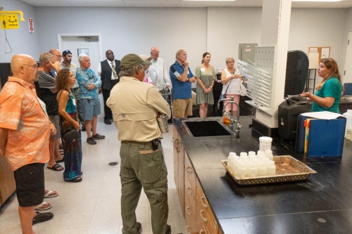 CanalWatch volunteers stand around a lab workstation as a City staff member explains water testing equipment, with sample bottles and instruments visible on the counter.